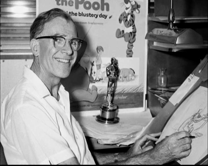 A black and white photo of Frank Thomas sitting at his drawing board smiling with Winnie the Pooh in the background