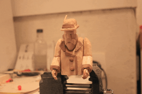 a man made out of wood with a long beard and a hat looking down at his hands and moving his hands around filmed in a stop motion animation style