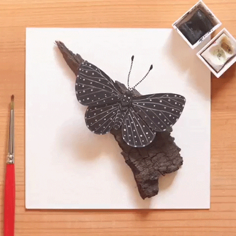 a black butterfly with white dots made from paper cut out animation fluttering its wing on a piece of bark which is sitting on a piece of paper on a desk
