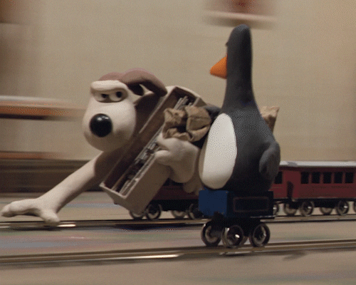 Grommet on a toy train racing a penguin on a small cart on tracks and he has a serious look on his face